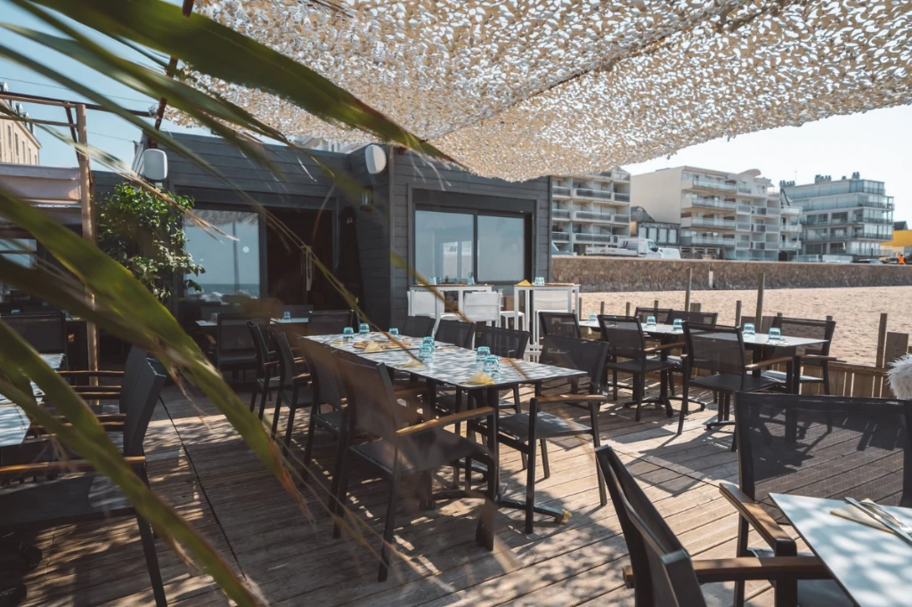 Terrasse de restaurant en bord de plage, tables et chaises sous un filet d'ombrage, devant un bâtiment au bardage gris