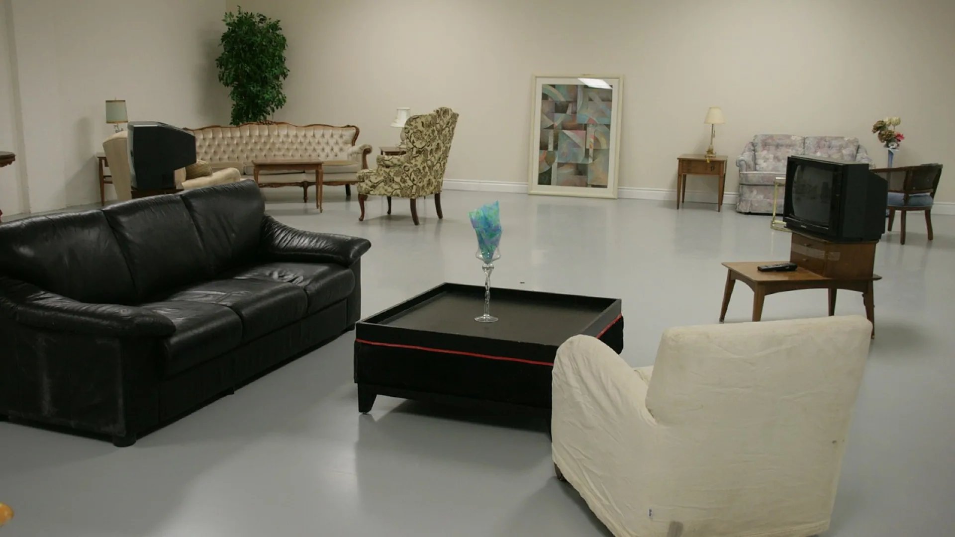 Grande salle au sol gris avec un canapé en cuir noir, une table basse noire, des fauteuils anciens et de vieux téléviseurs