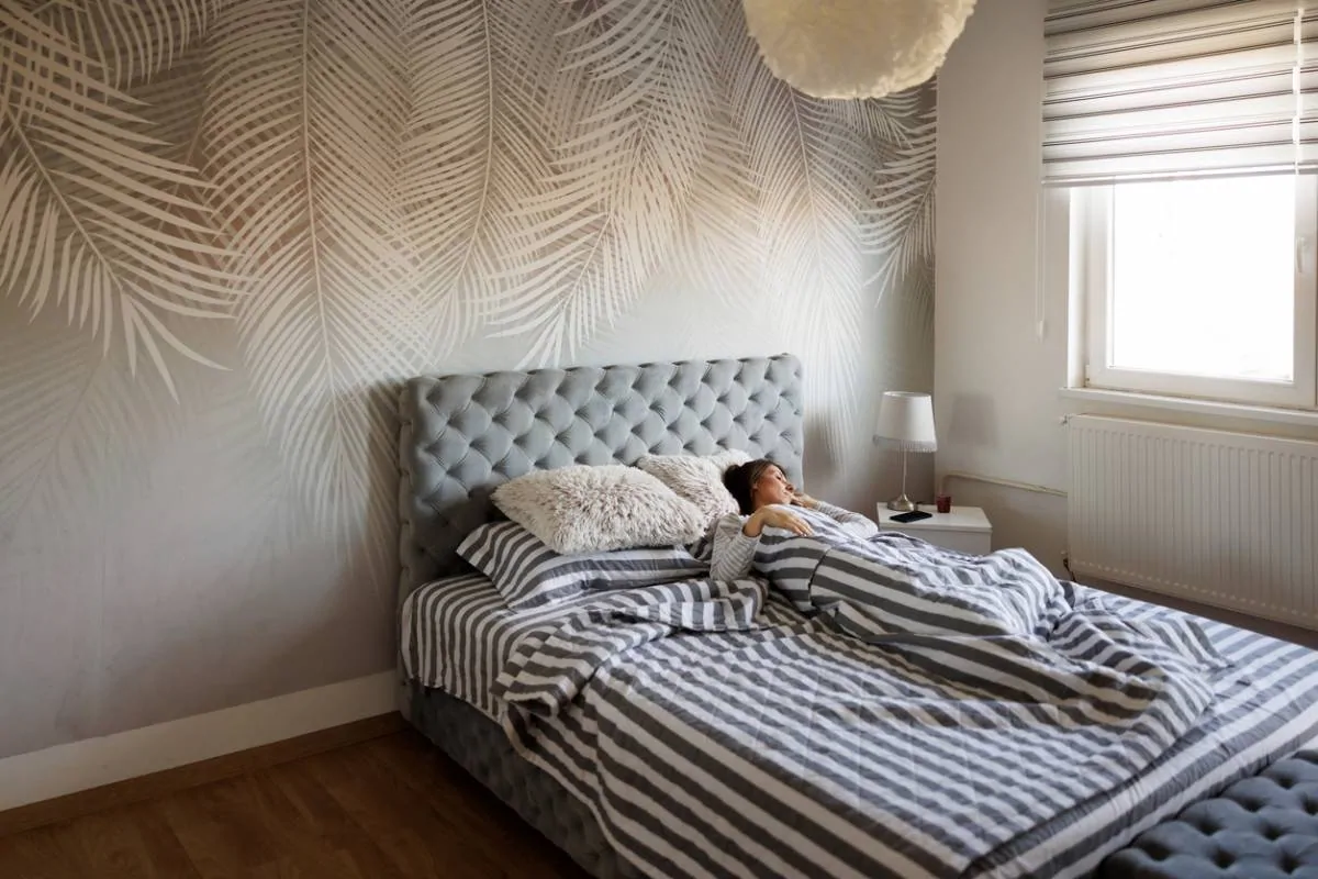 232447 Femme allongée dans un lit à tête capitonnée grise et parure rayée, devant un papier peint à motif de feuilles de palmier