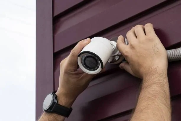 Mains installant une caméra de surveillance tube blanche sur un bardage bordeaux, près d'une gaine électrique