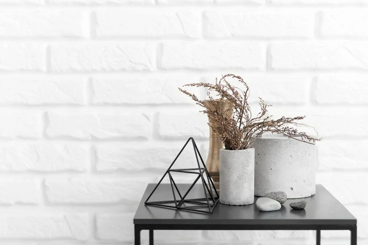 Console noire garnie de pots en béton, herbes séchées et pyramide en métal devant un mur de briques peintes en blanc