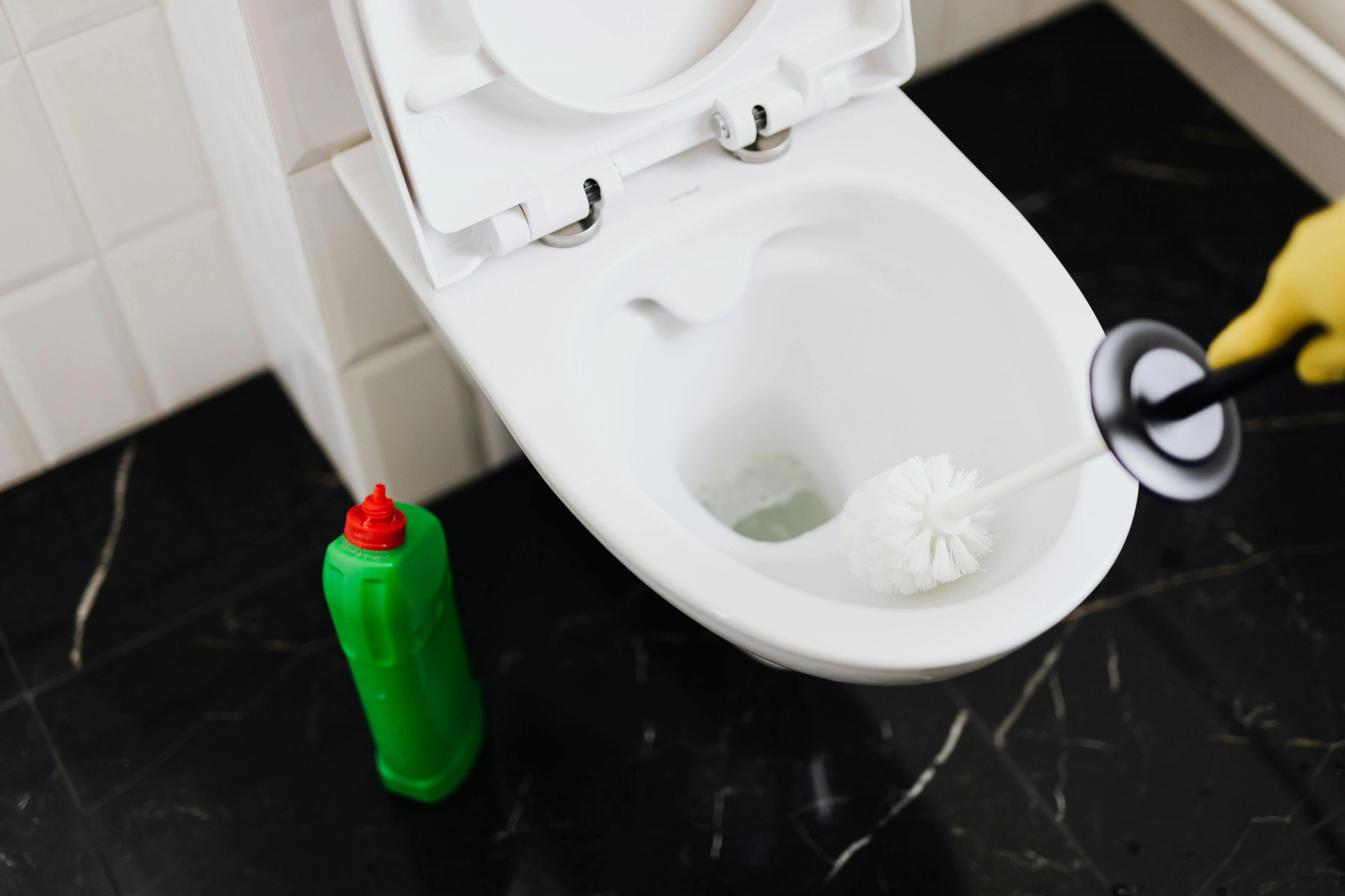 pexels-karola-g-4239086 Main gantée de jaune nettoyant une cuvette de WC suspendu à la brosse, flacon de gel vert posé sur le carrelage noir