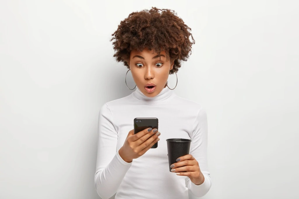 Jeune femme bouche bée devant l'écran de son smartphone, un gobelet de café noir dans l'autre main, sur fond blanc