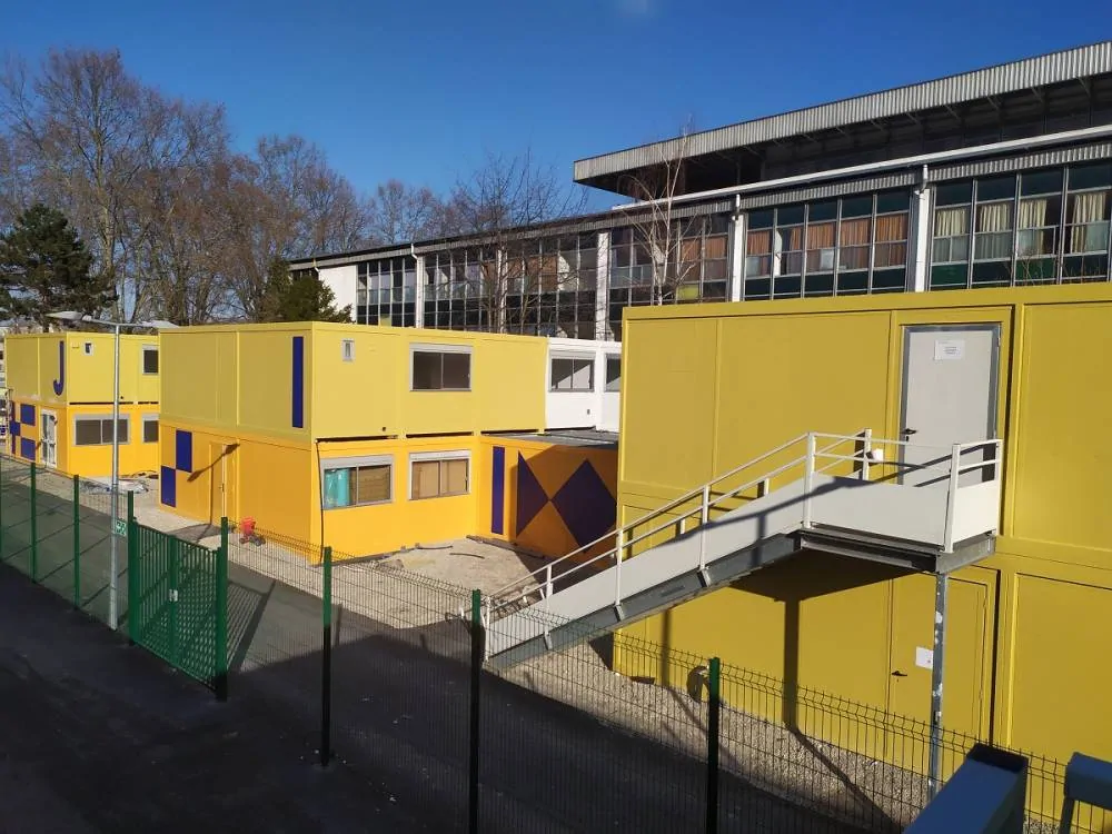 Bâtiments modulaires jaunes à bandes bleues reliés par un escalier métallique extérieur, au pied d'un grand bâtiment vitré